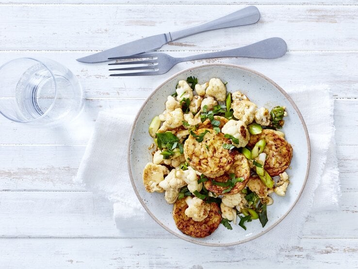 Gemüsepfanne mit Tempeh in einem hellblauen Teller auf einem Geschirrtuch neben einem Glas Wasser und Besteck auf einem weißen Tisch.
