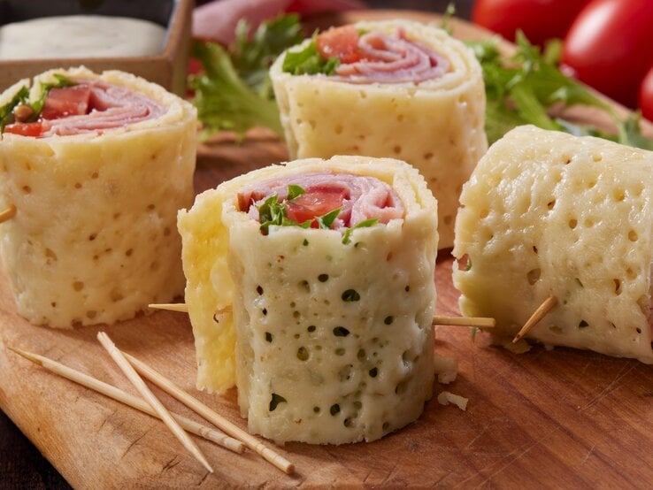 Mozzarella-Röllchen auf Holzbrett mit Zahnstochern. Im Hintergrund Salat, Tomaten und Dipschale. Frontalansicht.