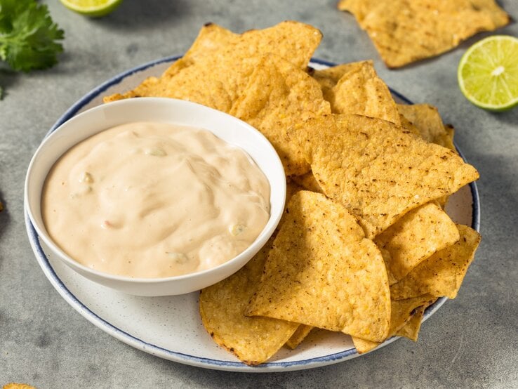 White Queso Dip mit Nachos auf weißem Teller und grauem Tisch. Im Hintergrund Limette, Nachos und Koriander. Draufsicht.