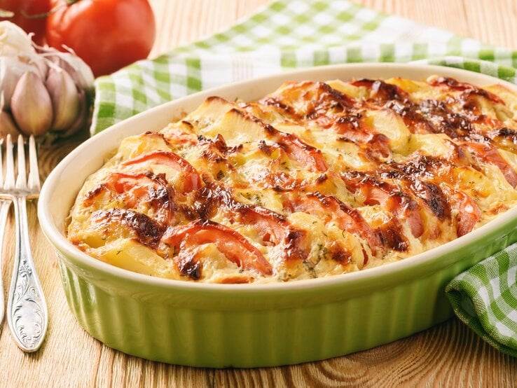 Eine Auflaufform mit Kürbis-Kartoffel-Auflauf mit Tomaten und Käse überbacken, daneben Tomaten, Knoblauch und eine Gabel.
