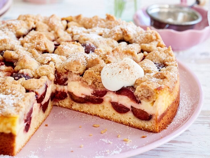 Pflaumen-Puddingkuchen mit Zimtstreuseln, angeschnitten und bestreut mit Puderzucker und serviert mit geschlagener, süßer Sahne auf einem rosa Tisch.  | © EAT CLUB / Isabell Triemer