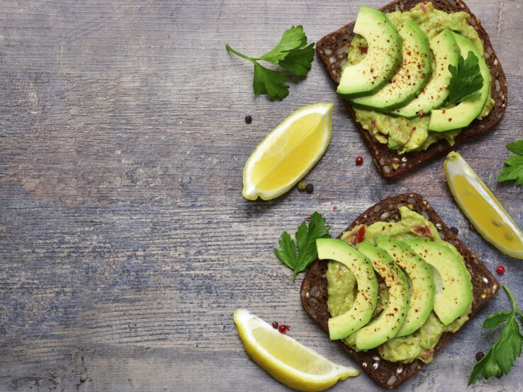 Zwei dunkle Brotscheiben mit Avocados belegt direkt von oben auf einem dunklen Tisch fotografiert.
