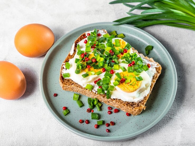 Draufsicht: Ein blauer Teller mit einem Hüttenkäse-Eier-Sandwich auf einem hellen Untergund. Daneben liegen zwei Eier mit Schale sowie grüne, schmale Blätter, wie von einer Lauchzwiebel.