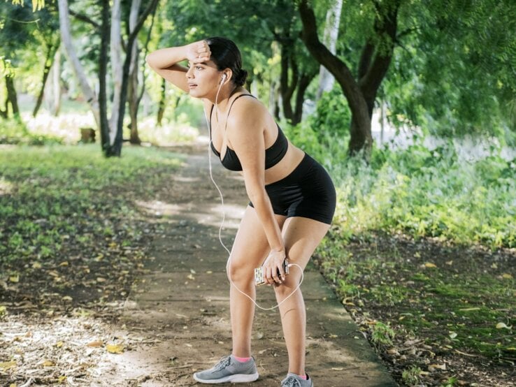 Eine Frau in Sportbekleidung macht eine Pause beim Joggen in einem grünen Park. Sie hält sich die Hand an die Stirn, hat Kopfhörer in den Ohren und sieht erschöpft aus.