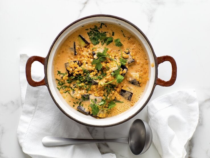 Rote Linsensuppe mit Auberginen und Kokosmilch in gusseisernem Topf garniert mit Koriander auf Marmorplatte; daneben auf Stoffserviette eine Suppenkelle