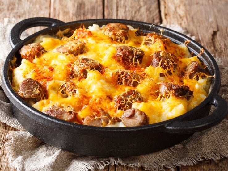 Draufsicht, schräg: auf Holzdielen steht eine runde und dunkle Auflaufform mit Griffen. Das Gericht heißt Jolean's Cheese Potato, es handelt sich um ein amerikanischen Auflauf mit Cabanissi-Würstchen.