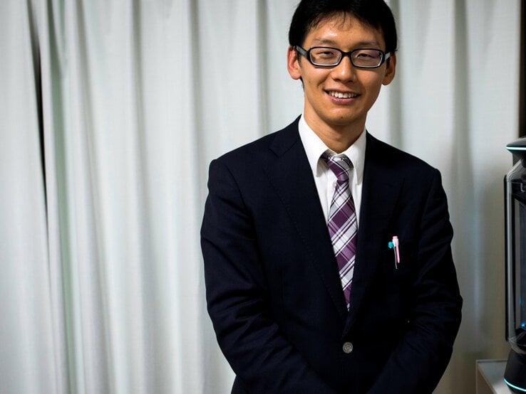 Mann mit kurzen dunklen Haaren und Brille trägt einen dunkelblauen Anzug mit einer lila-weiß gestreiften Krawatte und einem weißen Hemd. Er lächelt und steht vor einem weißen Vorhang.