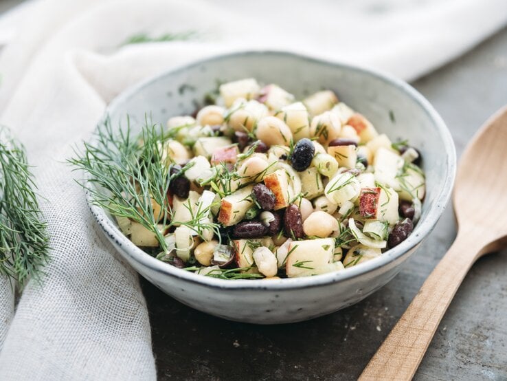 Eine Schüssel Apfel-Bohnensalat mit Kichererbsen und Dill, daneben ein Holzlöffel.