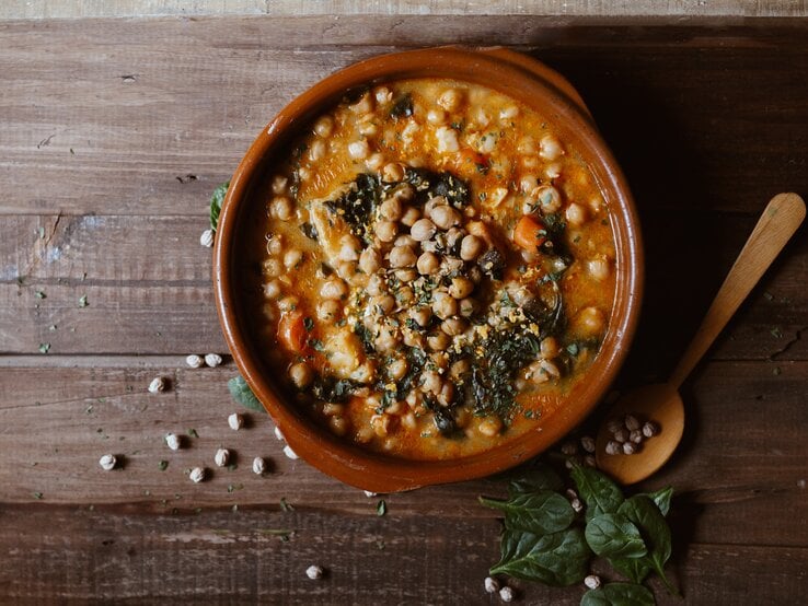 Eine Schale Mangold-Curry mit Kichererbsen in der Draufsicht, daneben ein Holzlöffel und ein paar Kichererbsen und Mangoldblätter.