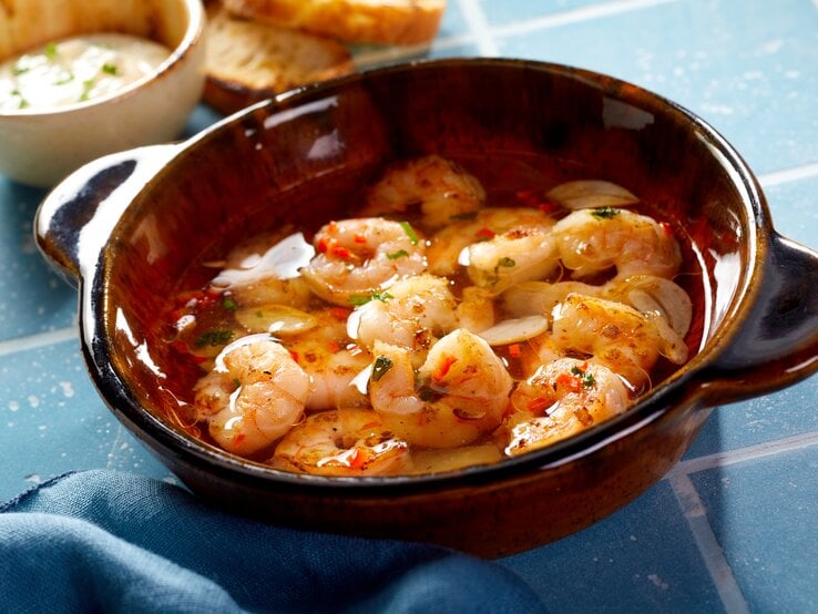 Draufsicht, schräg: Eine Tonschüssel mit Shrimps al ajillo steht auf blauen Kacheln mit Fugen. Daneben liegen Weißbrotscheiben und einen Schüssel mit einer hellen Creme.