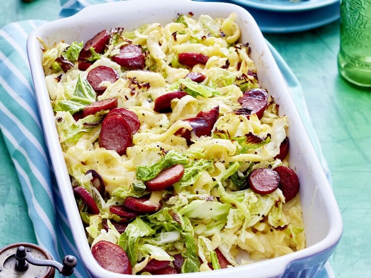 Auflaufform voller Wirsing-Käse-Spätzle mit angeschnittenen Würstchen, serviert auf einem hellen, blauenTisch. 