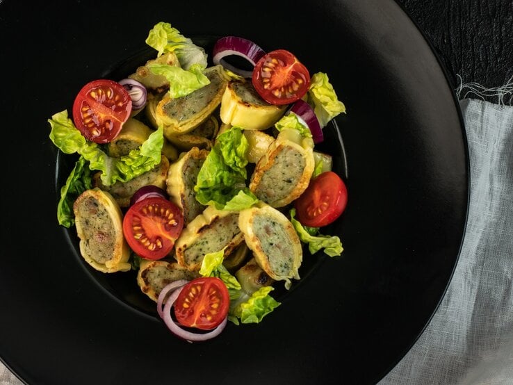 Ein schwarzer Teller mit dem Maultaschensalat auf einem dunklen Tisch mit hellgrauer Tischdecke. Direkt von oben fotografiert.