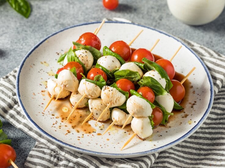 Fünf Tomaten-Mozzarella-Spieße liegen auf einem Teller. Neben dem Teller liegen einzelne Kirschtomaten und ein paar Blätter Basilikum.