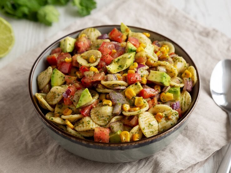 Eine Schüssel Nudelsalat mit Mais und Avocado, außerdem Paprika, drumherum frische Zutaten wie Kräuter und eine halbe Limette, außerdem 2 Löffel.