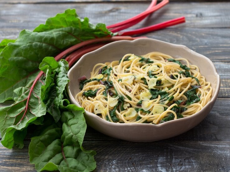Pasta mit Mangold in cremiger Frischkäse-Soße in beigem Teller auf Holztisch. Daneben bunter Mangold. Draufsicht.