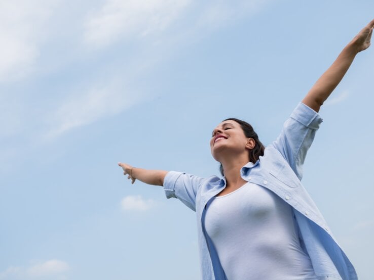  Das Bild zeigt eine Frau, die mit ausgebreiteten Armen nach oben steht und den Himmel anschaut. Sie scheint ein Gefühl von Freiheit oder Erleichterung zu erleben, vielleicht genießt sie einen Moment der Ruhe oder des Triumphes. Die Frau trägt eine blaue Bluse und ein weißes Top, und der Hintergrund besteht aus einem klaren blauen Himmel mit einigen leichten Wolken. Die Pose der Frau und ihr Gesichtsausdruck vermitteln ein starkes Gefühl der Freude und der Offenheit gegenüber der Welt um sie herum.