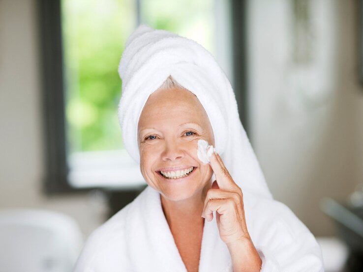 Lächelnde Frau mit einem Handtuch um ihren Kopf gewickelt und einem weißen Bademantel trägt Gesichtscreme auf ihre rechte Wange auf.  | © iStock/Tom Merton 