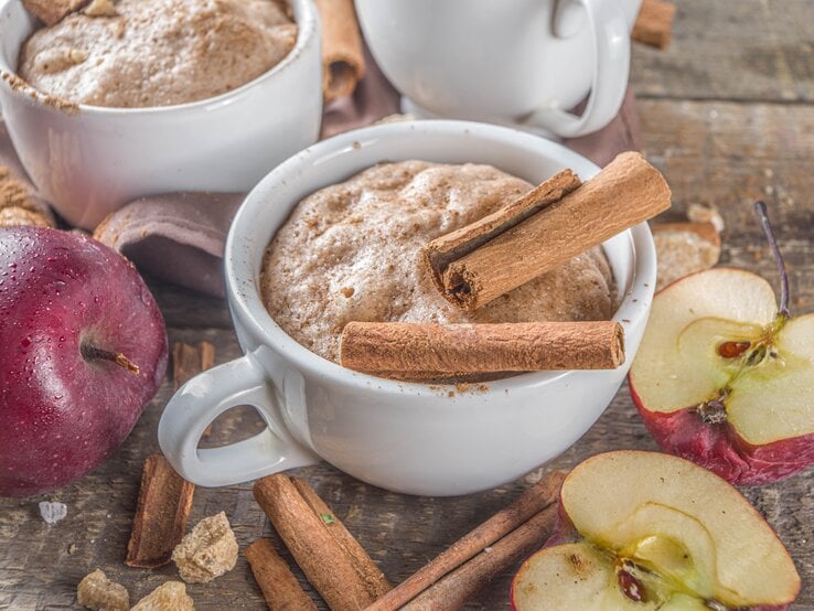 Zwei Tassen mit Apfel-Zimt-Tassenkuchen neben frischen Äpfeln auf einem Holztisch von schräg oben fotografiert.