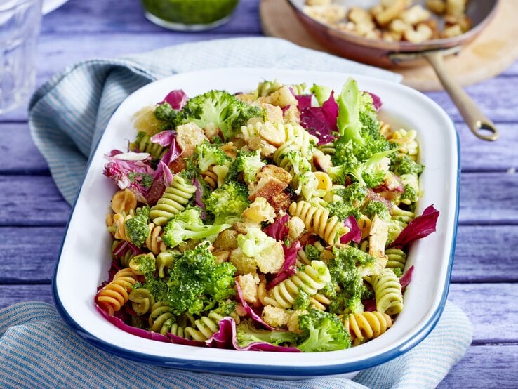 Eine weiße Auflaufform mit Brokkoli-Nudelsalat gefüllt auf einem violetten Tisch.