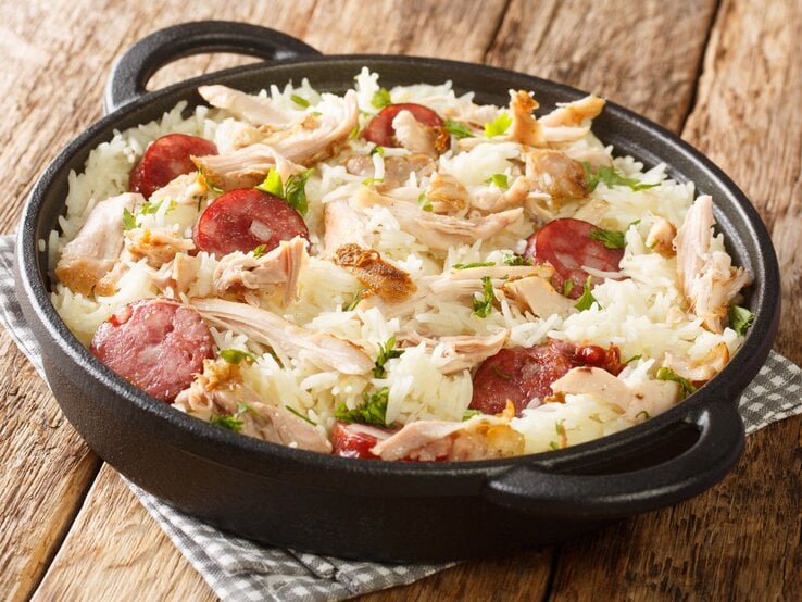 One Pot Reis mit Huhn und gebratener Wurst in Gusspfanne auf Stoffserviette auf Holztisch. Aufnahme aus der Draufsicht.