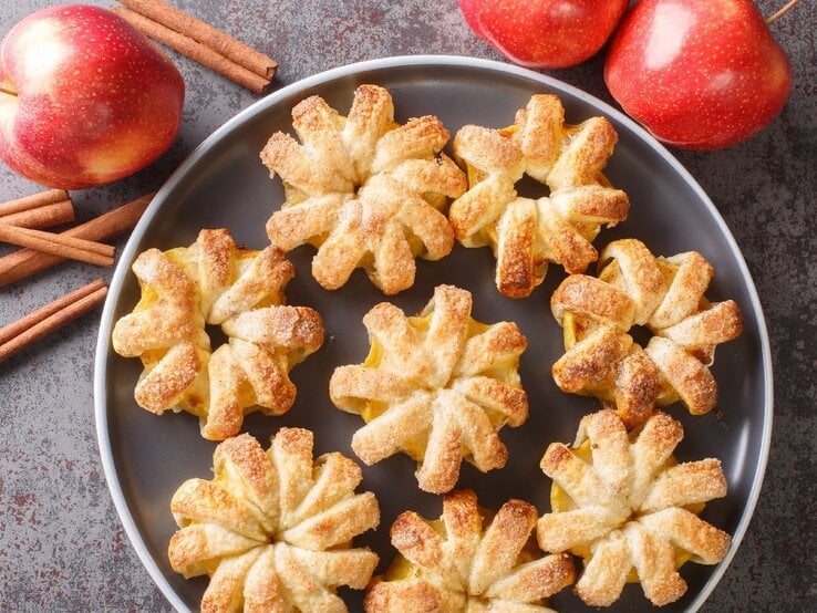 Ein grauer Teller mit den Apfelblumen auf grauem Untergrund. Daneben rote Äpfel und Zimtstangen. | © Shutterstock/ Sergii Koval