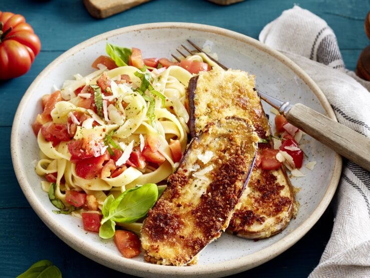 Auberginen-Schnitzel mit Bruschetta-Nudeln und Gabel auf einem hellen Teller. Drum herum Basilikum, Tomaten und ein Küchentuch, alles auf blauem Untergrund. 