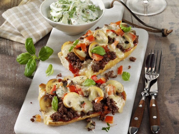 Französische Brotpizza mit Hackfleisch und Pilzen, garniert mit frischer Paprika und Basilikum und serviert mit einem frischen selbst gemachten Tzaziki auf einer weißen Steinplatte auf hölzernem Untergrund. 