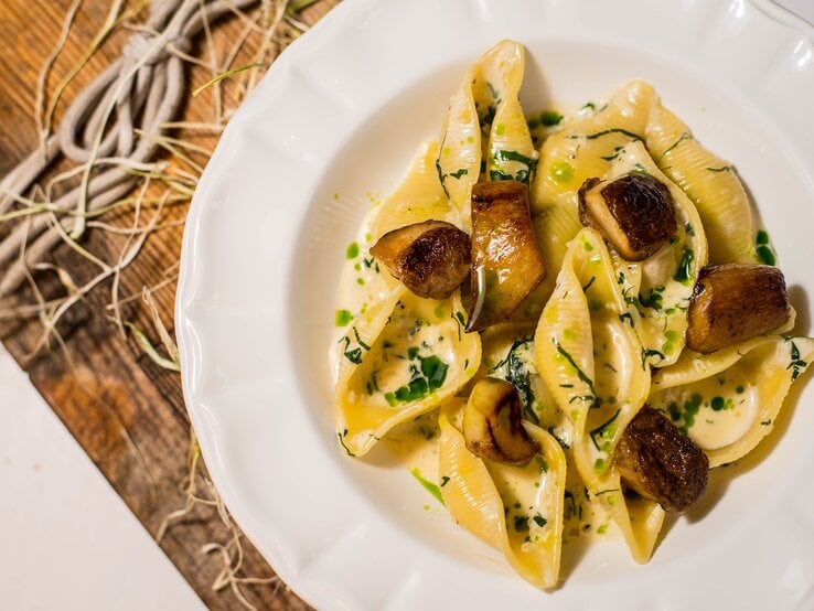 Draufsicht: Auf einem Holztisch steht ein weißer Teller mit cremigen Muschelnudeln mit frischen Pilzen