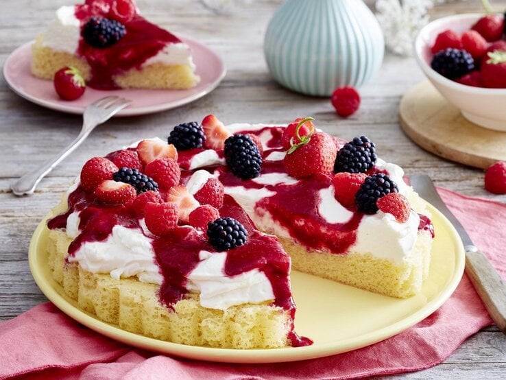 Auf einem Teller steht ein angeschnittener Schmand-Obstkuchen. Er ist mit Himbeersauce und Beeren überzogen. 
