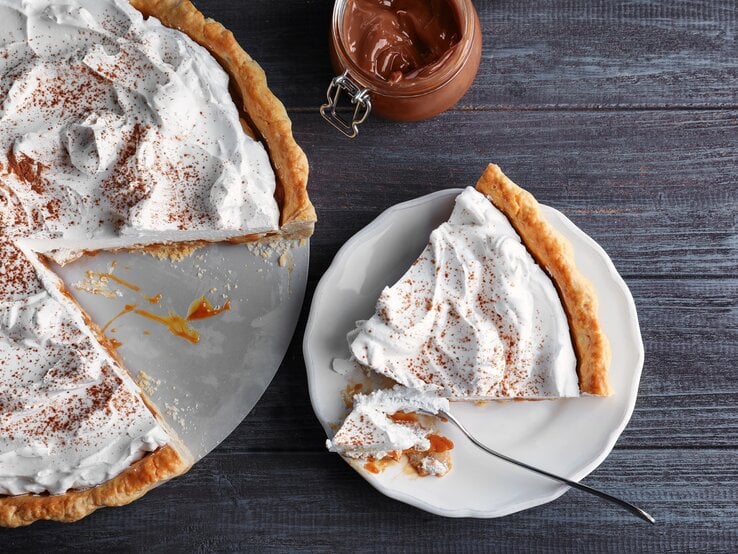 Ein angeschnittener Karamell-Kuchen mit Kokoscreme in der Draufsicht, daneben ein Weckglas mit Karamellsoße und ein Teller mit einem Stück des Kuchens.