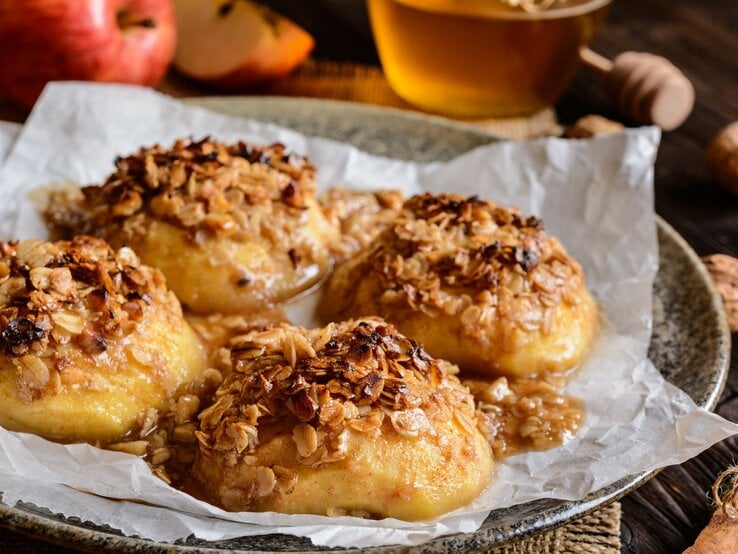 Vier gebackene Apfelhälften mit Haferflocken und Nüssen auf einem Teller mit Backpapier. Im Hintergrund unscharf die einzelnen Zutaten.