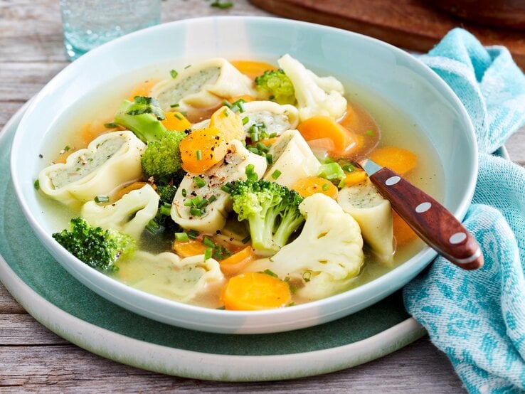 Weiße Schüssel mit Gemüsetopf mit Maultaschen und Löffel vor Holzhintergrund. 