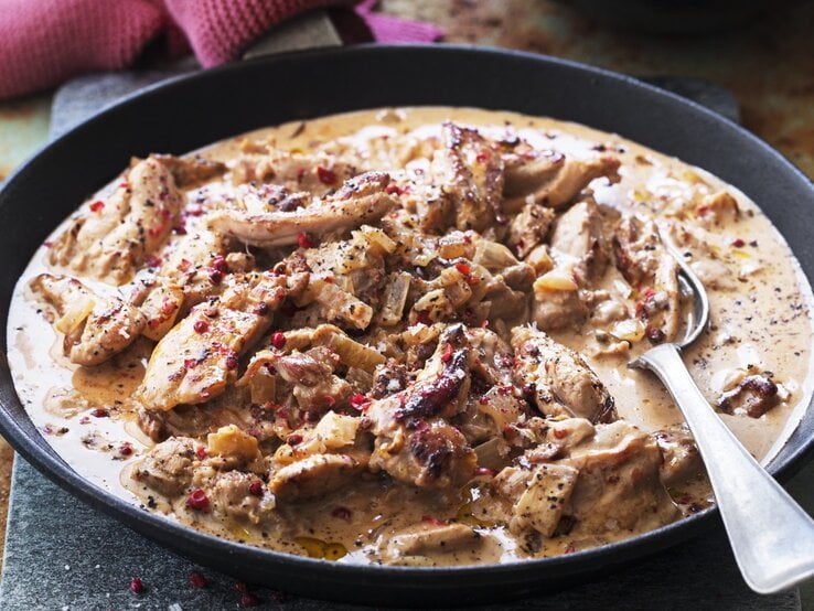 In einer tiefen Pfanne ist eine Portion Hühnereintopf mit Sahne und rosa Pfeffer angerichtet. Sie ist mit roten Pfeffer bestreut.