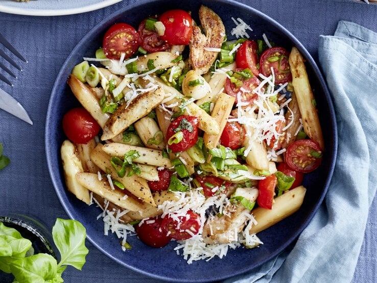 Italienischer Spargel-Mix mit Parmesan in dunkler Schüssel auf dunkelblauem Untergrund. Daneben Küchentuch, Besteck und Basilikumpflanze. 