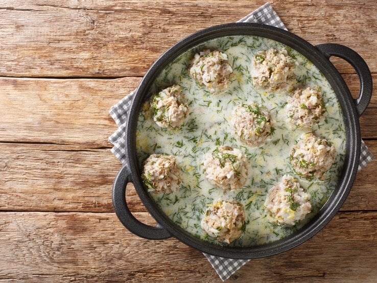 Saftige Reis-Hackbällchen serviert in cremiger Dill-Sahne-Soße in einer schwarzen gusseisernen Form. Das Ganze steht auf einem mit einem karierten Tischtuch bedecktem Holztisch.