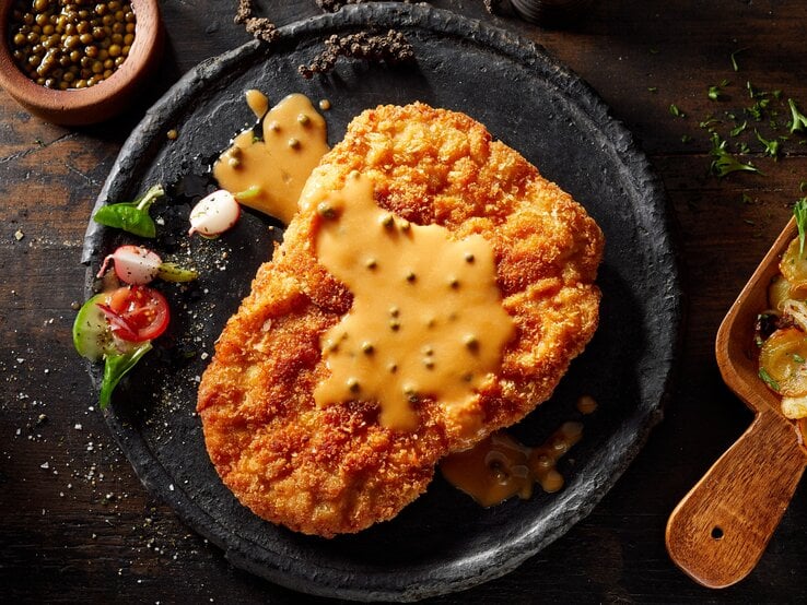 Ein Schwarzer Teller mit dem Schnitzel und der Senfsoße auf einem rustikalem Untergrund.