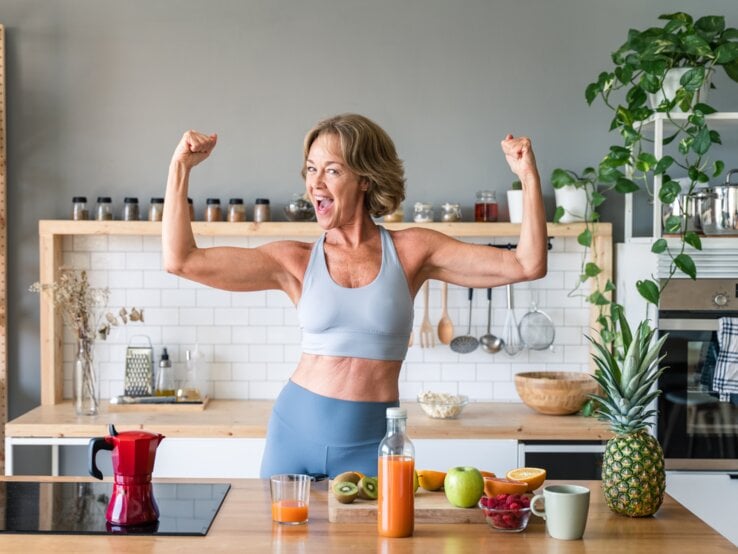 Eine ältere Frau, die Sportbekleidung trägt, steht in einer Küche und hat die Arme angewinkelt, sodass sie ihre definierten Arme präsentiert. Vor ihr auf dem Tisch liegt etwas Obst sowie eine Saftflasche.