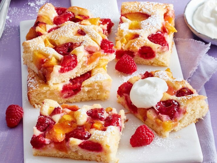 Auf einem Tisch sind mehrere Stücken Pfirsich-Melba-Kuchen serviert. Sie sind mit Puderzucker bestreut.