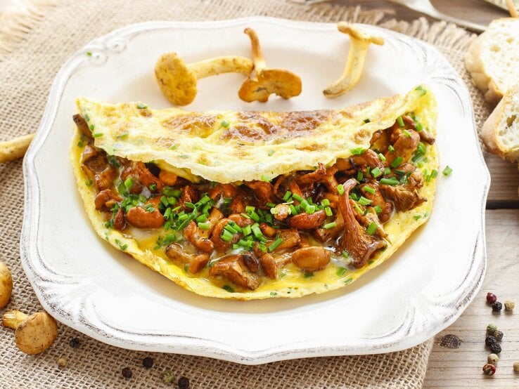Auf einem weißen Teller liegt ein Pfifferling-Omelette. Um den Teller herum liegen frische Pfifferlinge, Pfefferkörnern und Weißbrotscheiben.