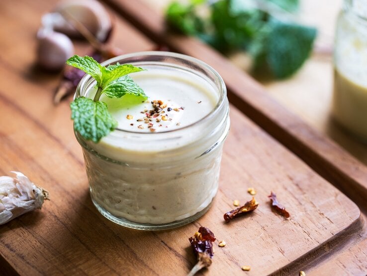 Ein Glas weißes Knoblauch-Zitronen-Dressing mit Minze und Pfeffer dekoriert, drumherum frische Zutaten.