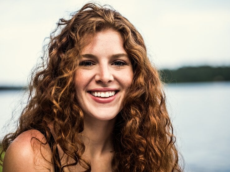 Junge Frau mit langen, lockigen roten Haaren, die in die Kamera lächelt. Sie hat einen frischen, natürlichen Look und strahlt Freude und Zufriedenheit aus.