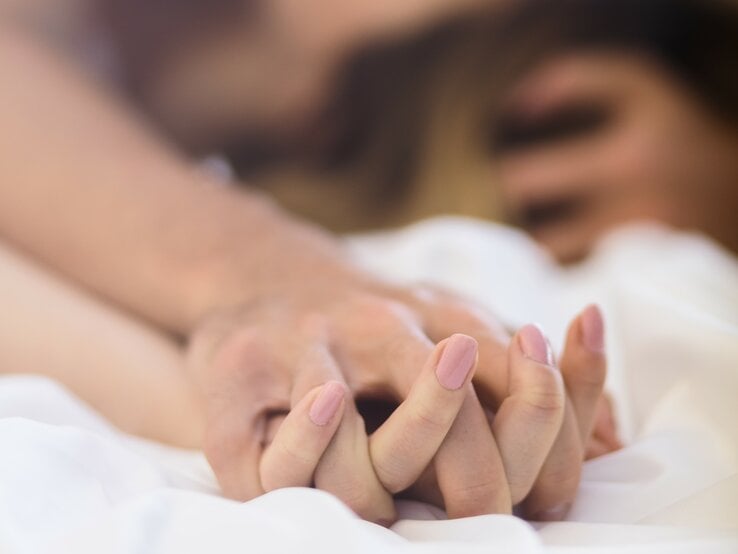 Hände eines Paares, die sich festhalten, im Vordergrund, mit unscharfen Gesichtern im Hintergrund, was Intimität andeutet.