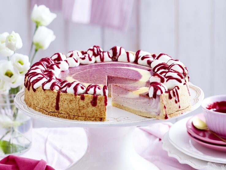 Auf einem Tortenteller steht ein beeriger Zebra-Käsekuchen. Er ist mit Sahnehäubchen und Soße verziert. | © EAT CLUB / Isabell Triemer