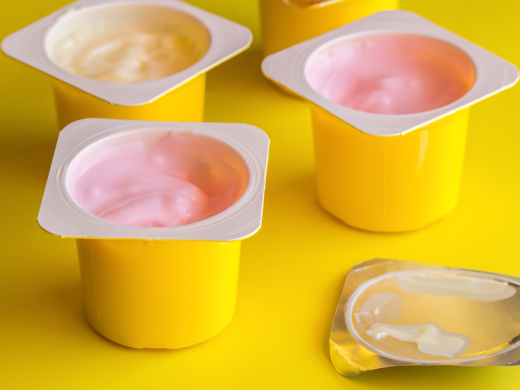 Fruchtaromatisierter Jogurt in gelben Plastikbechern auf hellgelbem Hintergrund mit silbernem Foliendeckel - Yogurt-Cup-Hintergrund mit selektivem Fokus