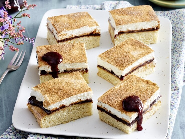 Sechs Pflaumen-Schmand-Schnitten auf weißem Teller, darunter ein blau geblümtes Küchentuch und blauer Untergrund, daneben Gabel und Blume.  | © EAT CLUB / Isabell Triemer 