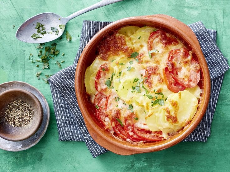 Tomaten-Kartoffel-Auflauf, überbacklen mit Käse und bestreut mit frischen Kräutern. Serviert in einer großen, runden Auflaufform auf einem türkisenen Untergrund. 