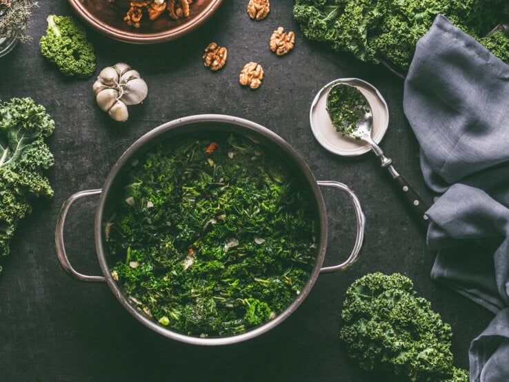 Ein Topf Grünkohl mit Räuchertofu in der Draufsicht, drumherum liegen Kohlblätter und viele weitere Zutaten.