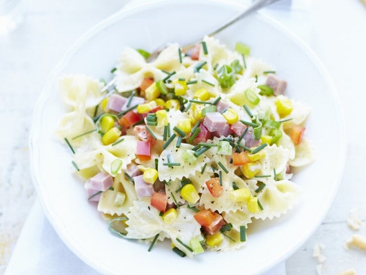 In einer Schüssel ist eine Portion Nudelsalat mit Farfalle, Schinkenwürfeln und Mais angerichtet.