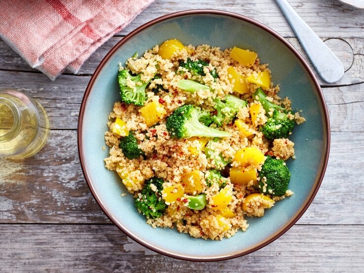 Brokkoli-Bowl mit Couscous in blauer Schüssel auf Holzuntergrund. Daneben rotes Küchentuch, Glasbehälter und das Ende einer Gabel/eines Löffels. 