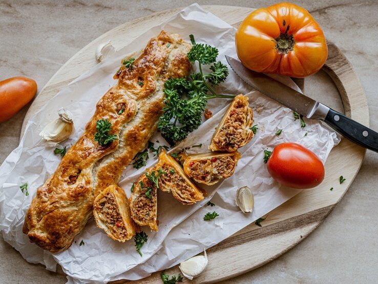 Hackfleisch-Blätterteig-Rolle auf einem Holzbrett. Daneben ein Messer und mehrere Tomaten.  | © Shutterstock/Angelika Heine